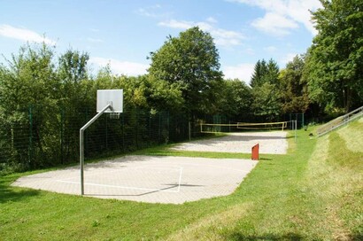 gepflasterter Platz mit Basketballkorb, im Hintergrund Sandplatz mit gespannten Netzen, gesäumt von grüner Wiese und grünbelaubten Bäumen