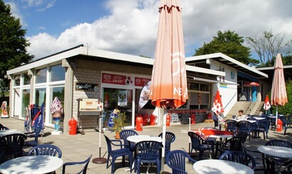 Außenbereich, Sonnenschirme geschlossen, weiße Tische, blaue Stühle, Kiosk im Hintergrund 