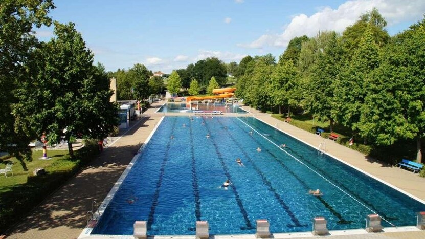 Blick auf das Schwimmerbecken, gesäumt von Baumbestand, im Hintergrund Rutsche im Nichtschwimmerbecken