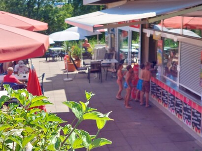 Freibadkiosk mit Außenbereich, helle Markise, rote Sonnenschirme, Kinder stehen am geöffneten Kioskfenster