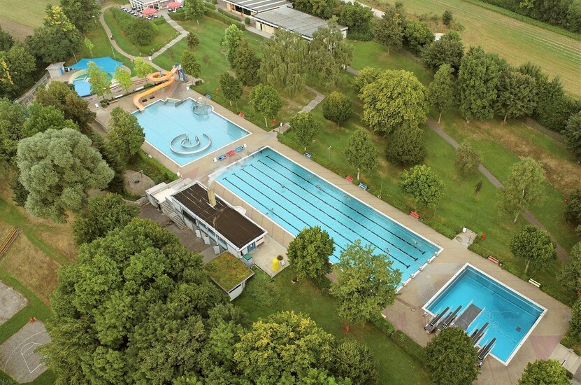 Schwimmbadgelände Ansicht von oben mit allen Becken, Gebäuden, Liegewiese mit Baumbestand